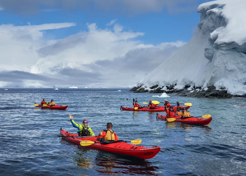 Kayakers greeting the camera beneath snow-covered Antarctic cliffs